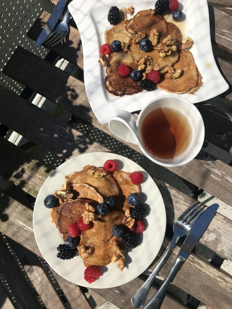 two plates of stacks of vegan banana pancakes topped with berries and nuts plus a cup of tea
