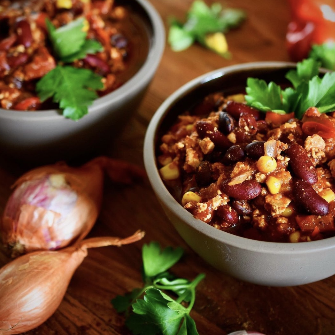 a vegan chili with beans, pepper, and corn in a bowl on a table decorated with shallots and parsley