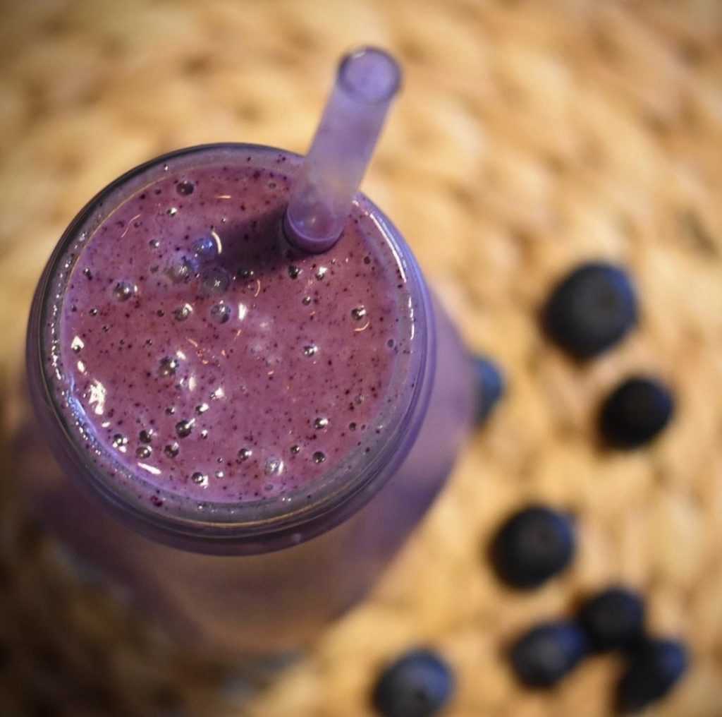 A dark purple blueberry smoothie in a glass with a straw against the backdrop of fresh blueberries