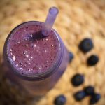 A dark purple blueberry smoothie in a glass with a straw against the backdrop of fresh blueberries