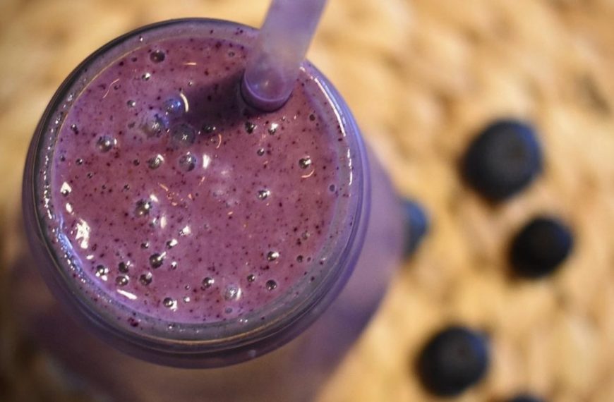 A dark purple blueberry smoothie in a glass with a straw against the backdrop of fresh blueberries