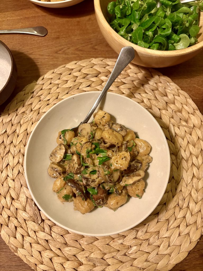 a plate of gnocchi with mushrooms and parsley and some salad in the background