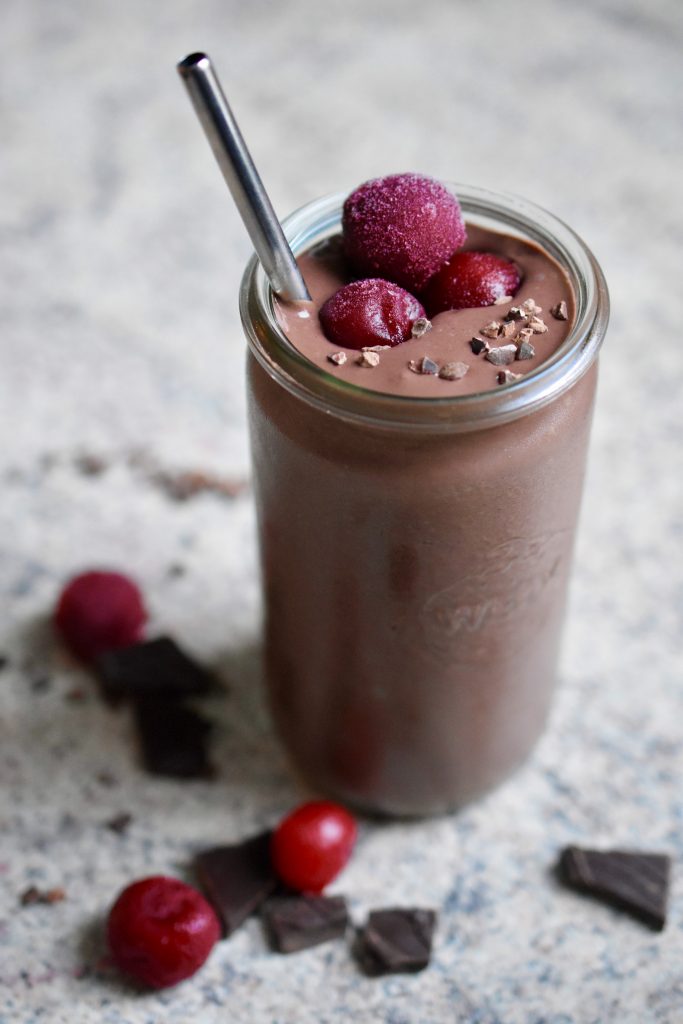 a thick, creamy vegan chocolate cherry smoothie decorated with frozen cherries and cacao nibs in a glass with a metal straw