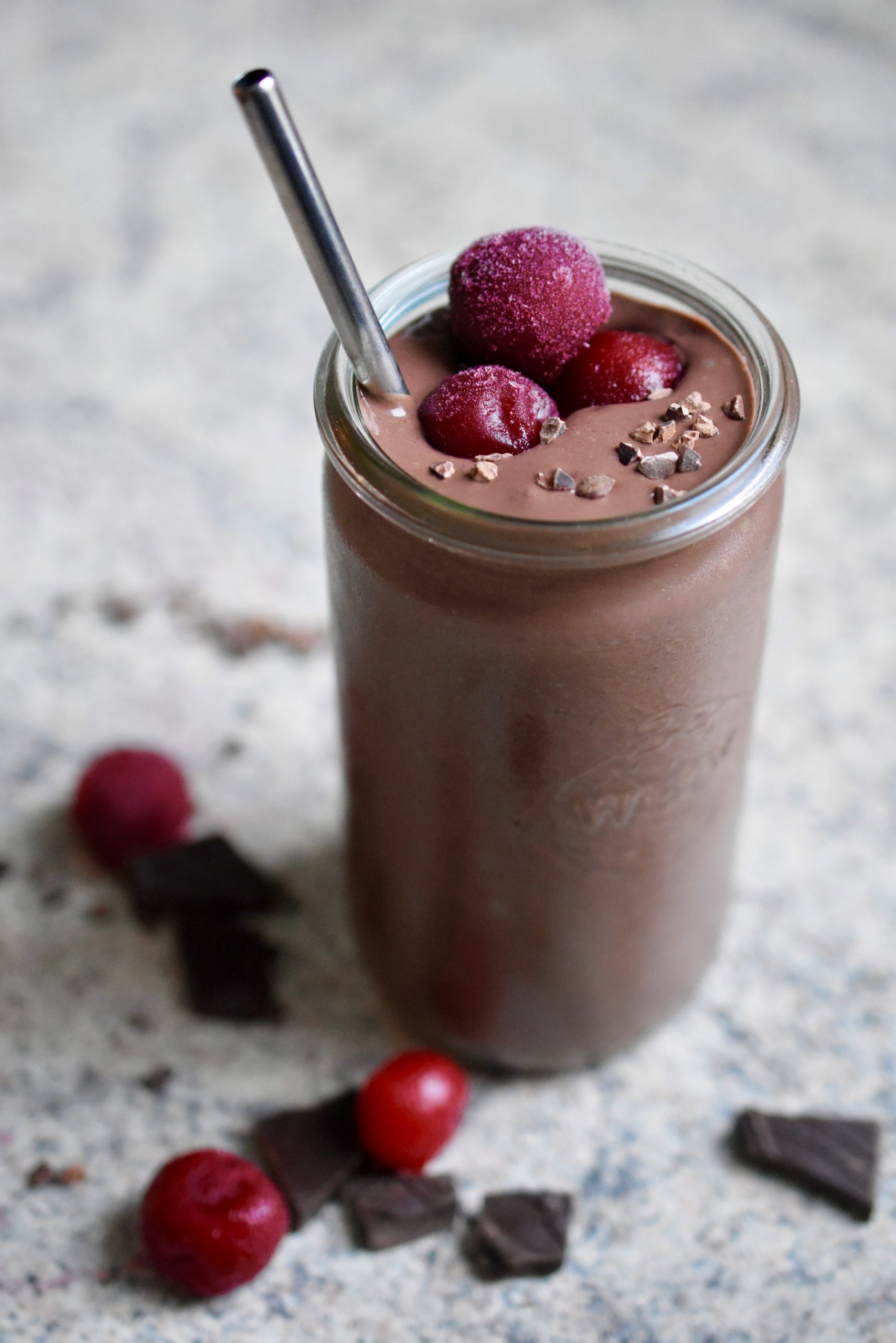 a thick, creamy vegan chocolate cherry smoothie decorated with frozen cherries and cacao nibs in a glass with a metal straw